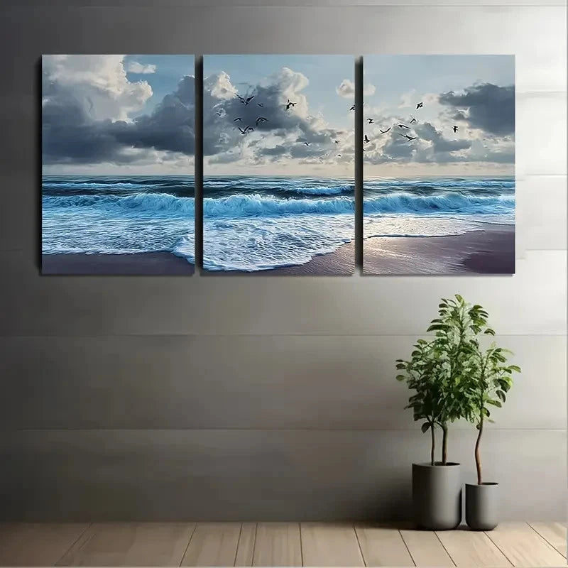 Triptych wall art of a beach scene with waves and clouds, displayed on a wall with two potted plants below.