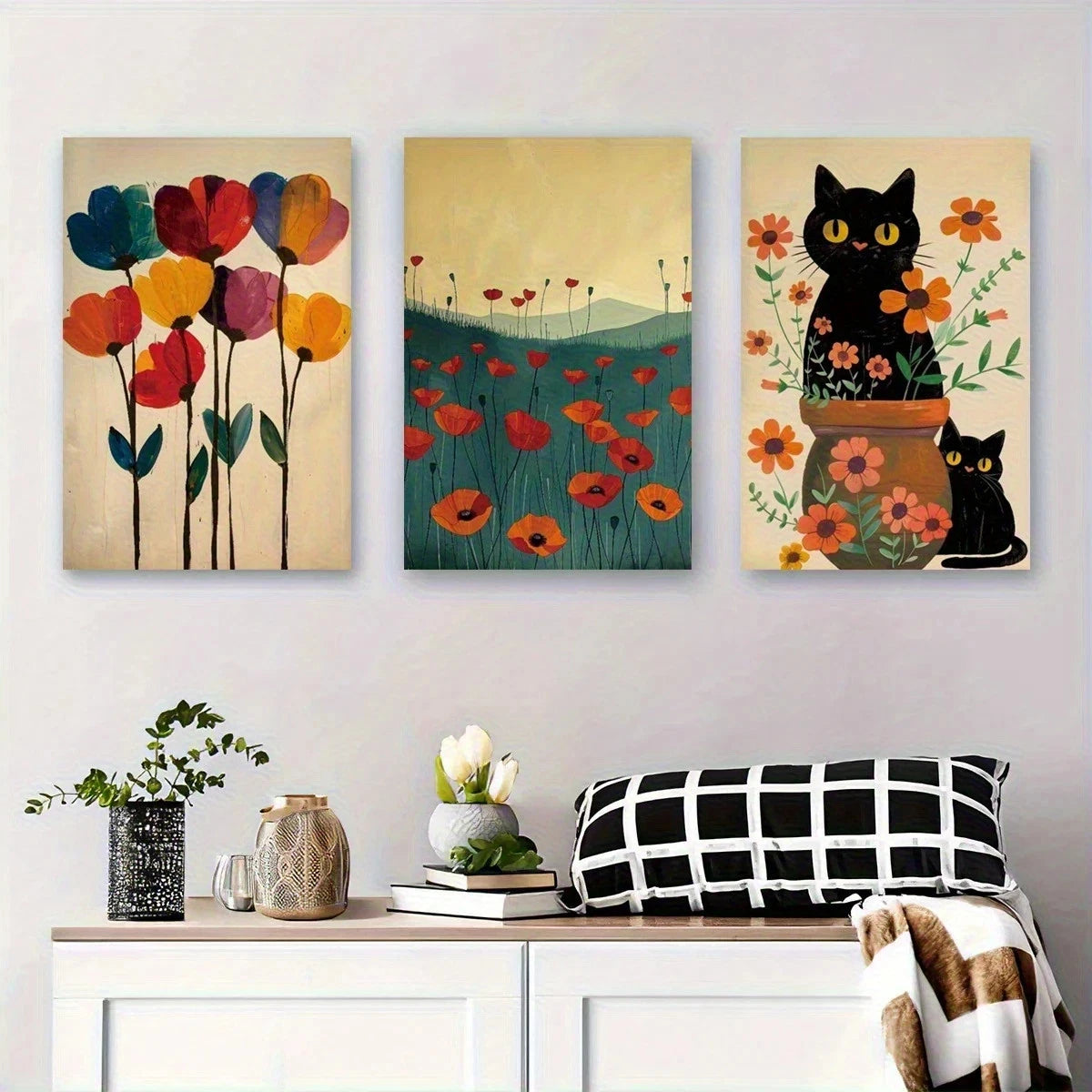 Three colorful paintings of flowers and cats on a wall above a white cabinet with decorative items.