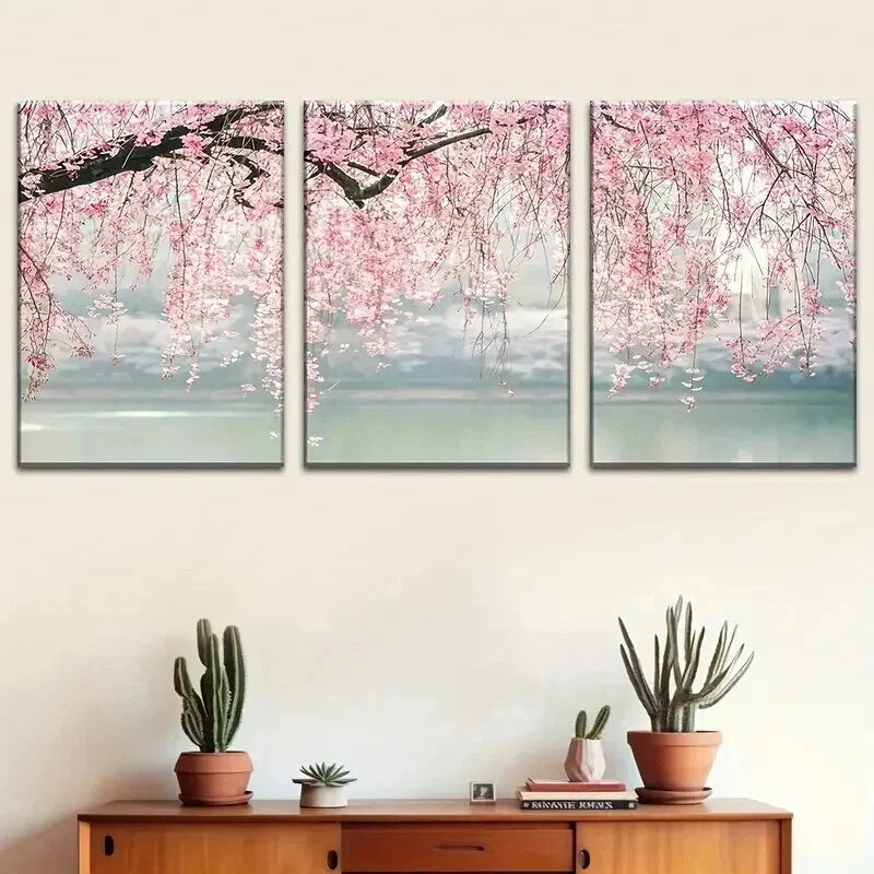 Three-panel wall art of cherry blossom trees over water with a wooden cabinet and plants below.