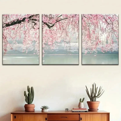 Three-panel wall art of cherry blossom trees over water with a wooden cabinet and plants below.